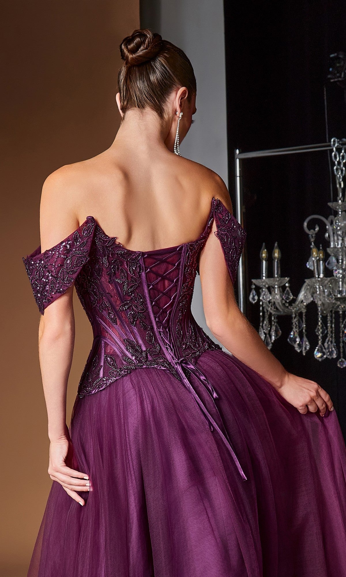 A woman in the Ladivine CD657 long prom dress, featuring an off-the-shoulder deep purple design with a corset bodice, lace-up back, and beaded tulle skirt, stands facing away while holding the embellished gown.